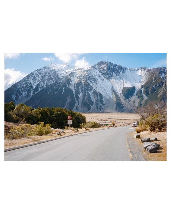 A winding road leads towards snow-capped mountains under a blue sky with scattered clouds.