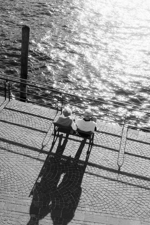 A couple sits on a bench by a body of water with sunlight reflecting off the surface.