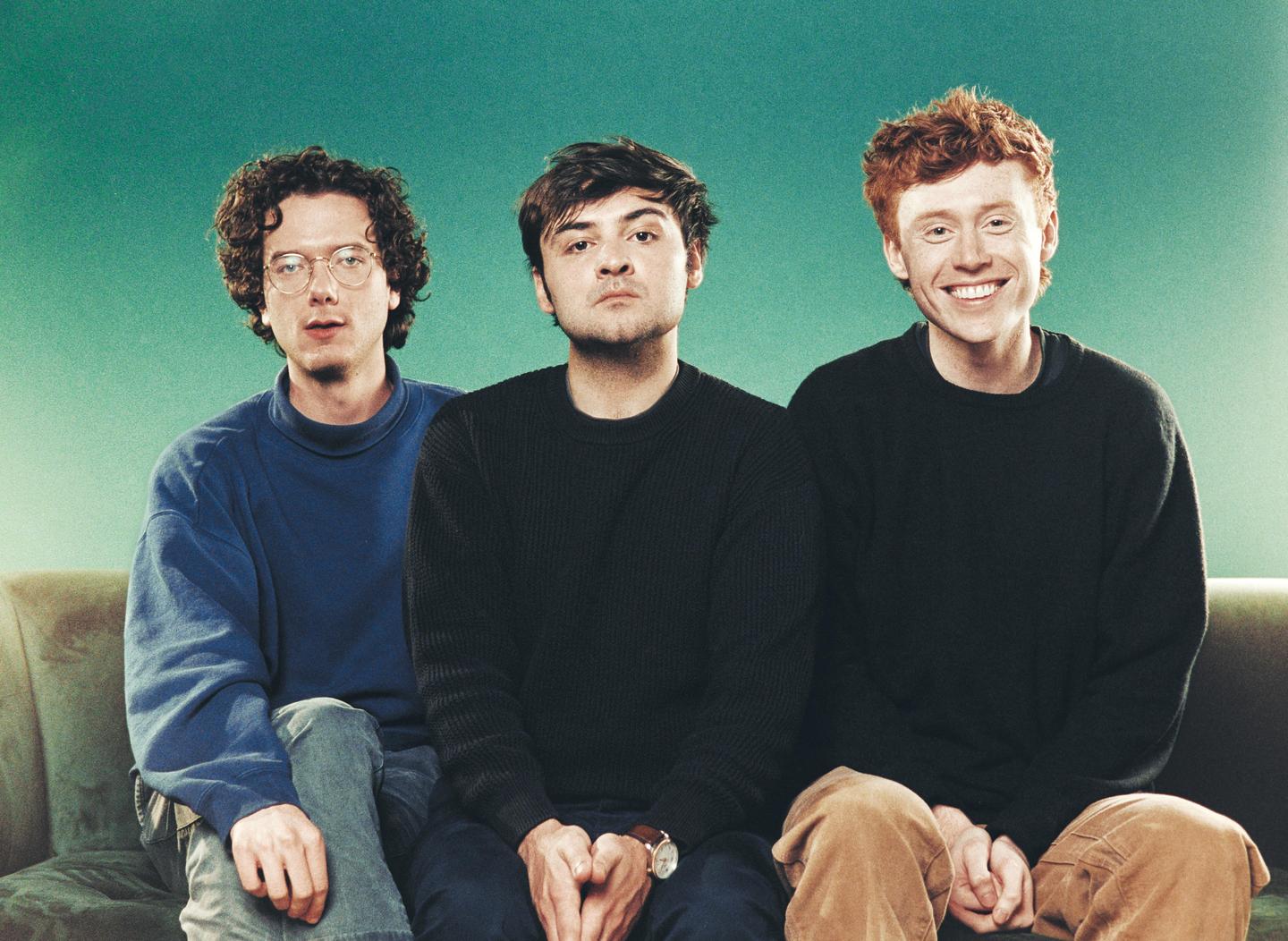 Three men are sitting on a couch in front of a teal background.
