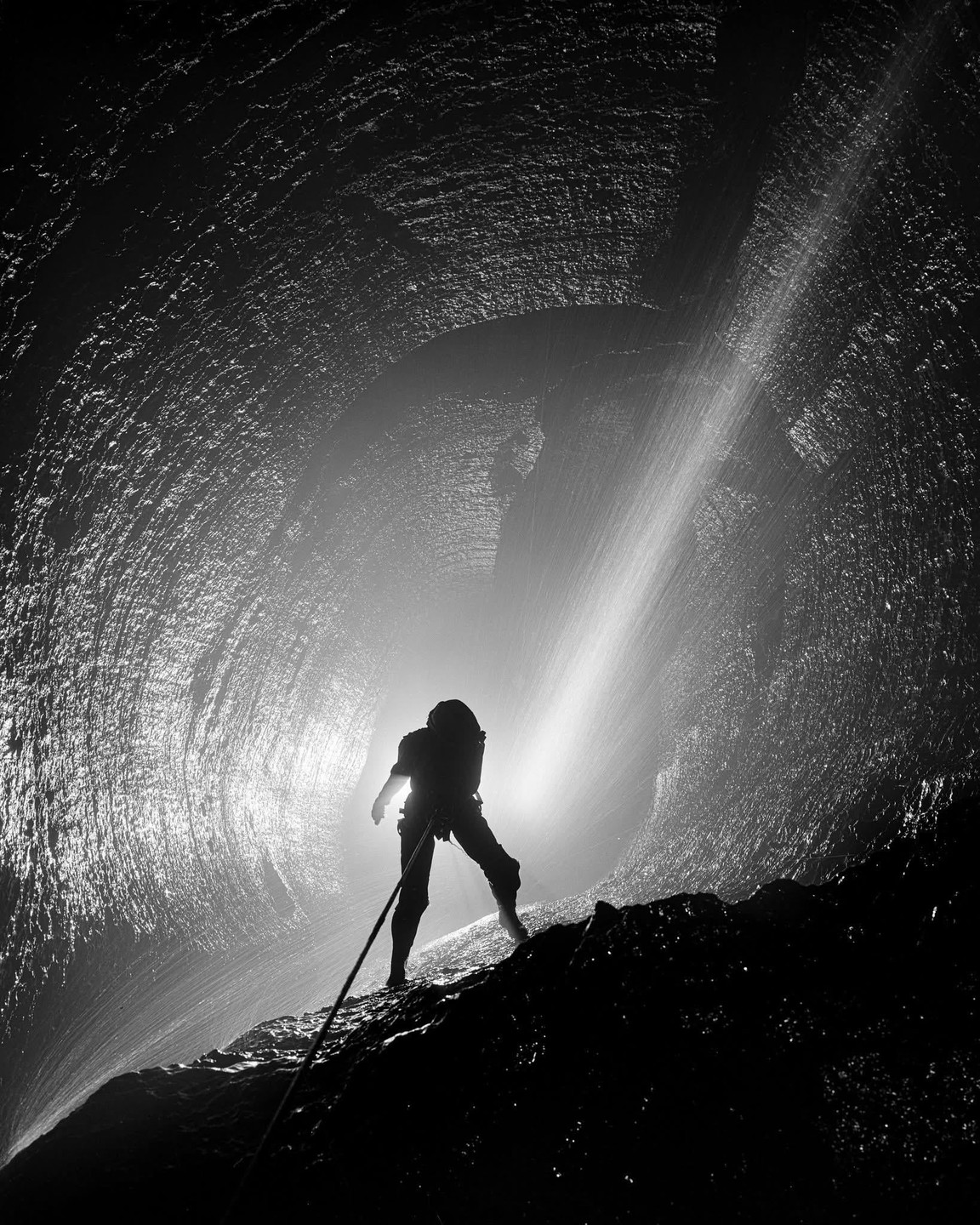 A person rappels down a rock face with a waterfall and bright light in the background.