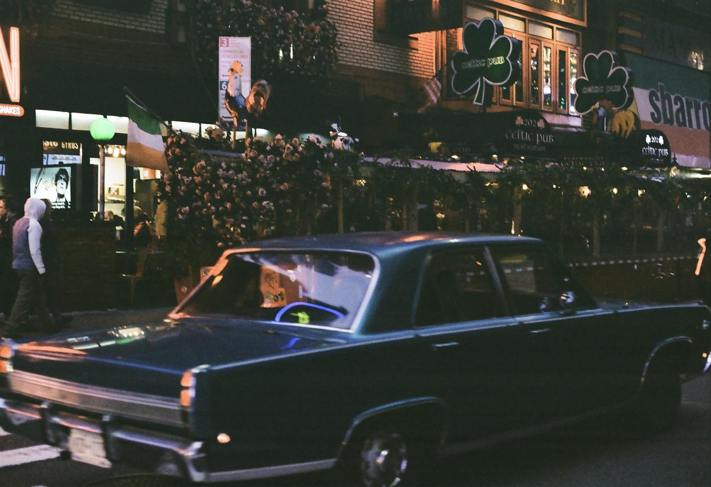 A dark blue car is parked on a street at night in front of a pub with shamrock signs.