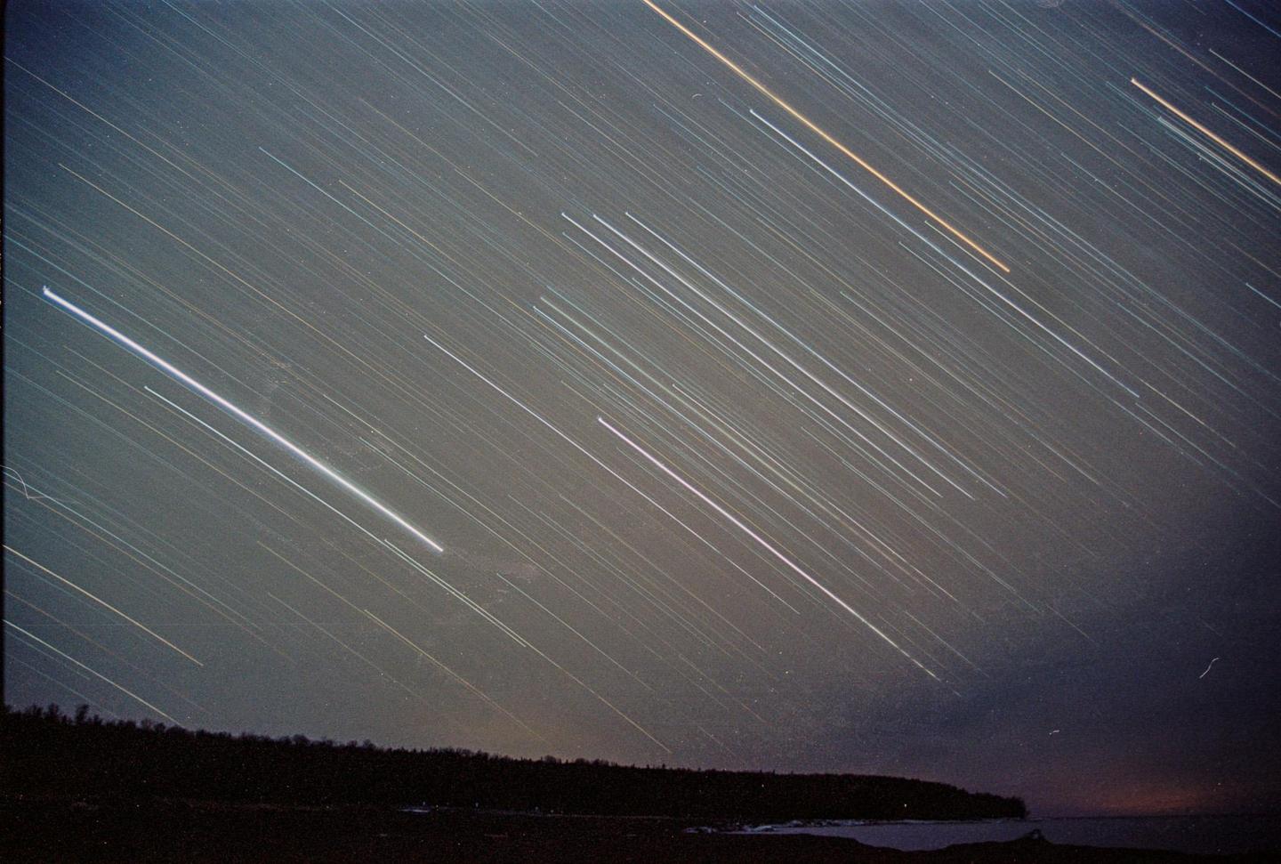 Star trails streak across the night sky above a dark shoreline with a sliver of water visible.