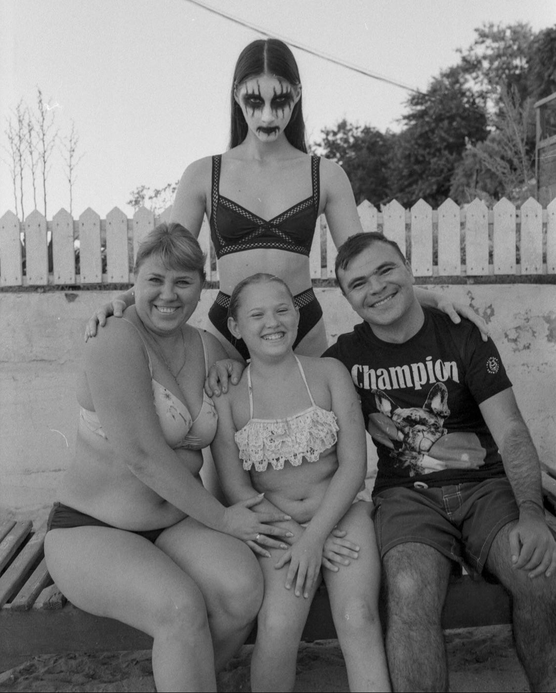 A woman with dark makeup stands behind a family of three sitting on a bench.