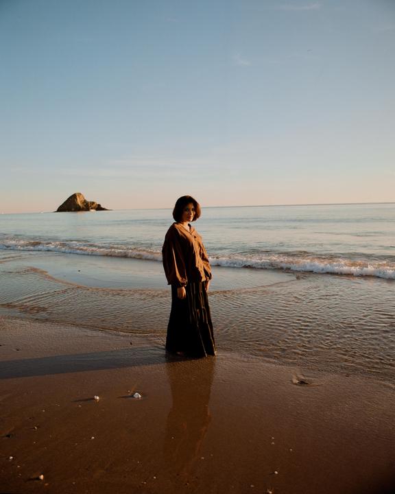 A woman stands in the shallow water of a beach with a rocky island in the distance under a clear sky.