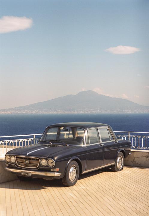 A dark blue vintage car is parked on a wooden deck overlooking a body of water with a mountain in the distance.