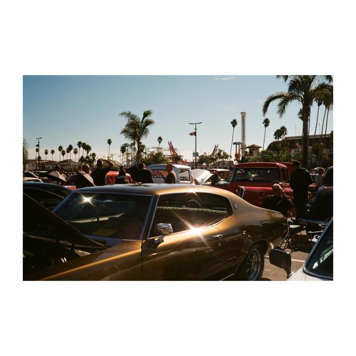 A gold vintage car is parked among other vehicles and people under palm trees with amusement park rides in the background.