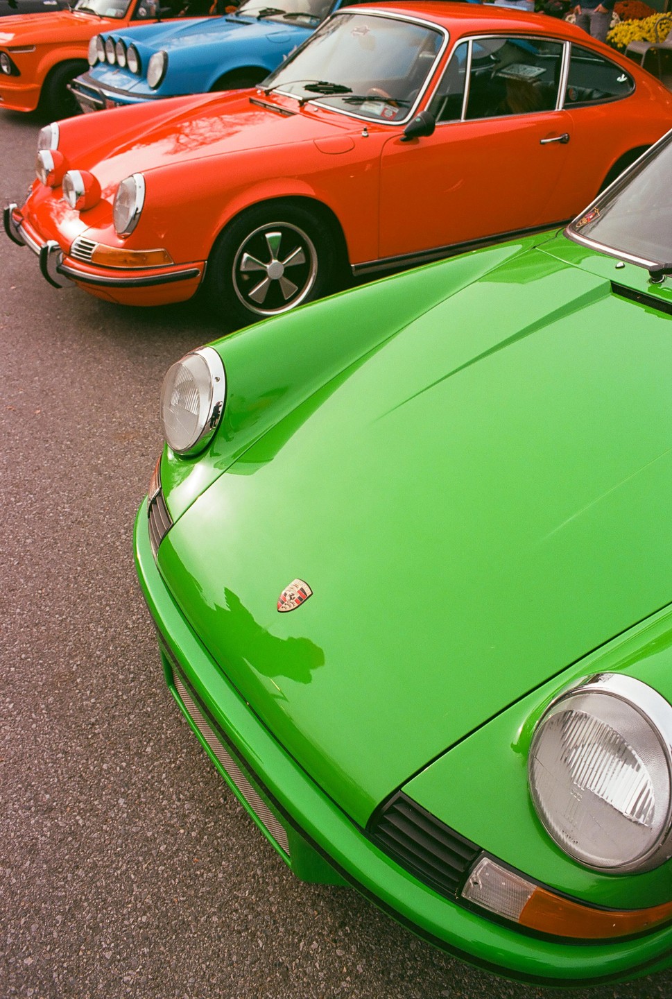 Three vintage cars, green, orange, and blue, are parked in a row on asphalt.