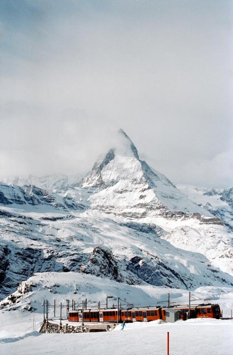 An orange train is visible at the base of a snow-covered mountain with a cloudy sky.