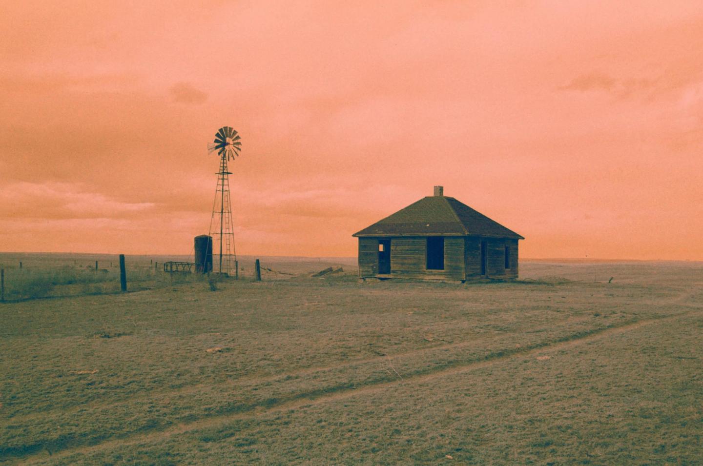 An abandoned house and a windmill stand in a vast, grassy field under an orange sky.