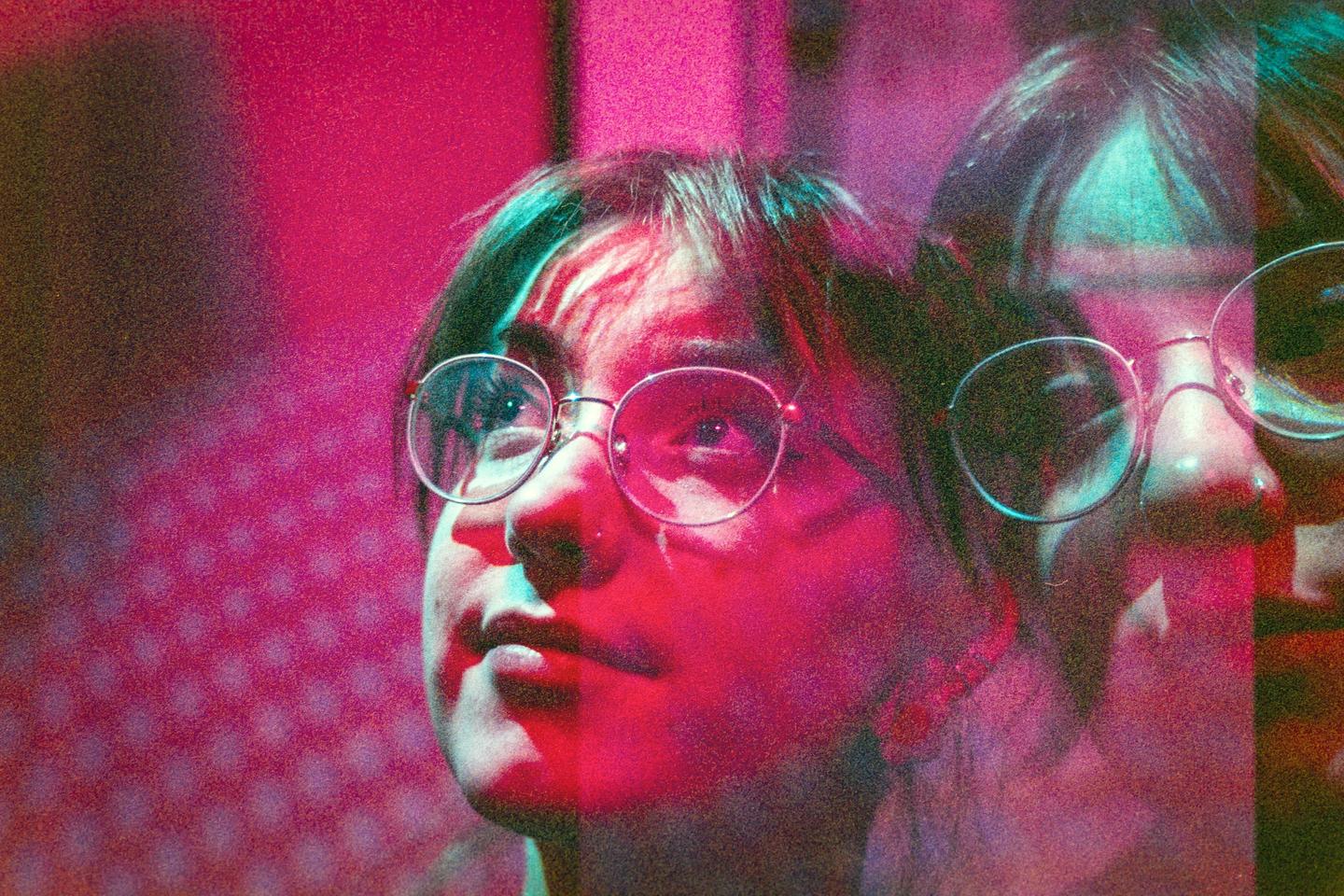 A woman with glasses looks up, illuminated by pink and blue light, with a faint double exposure of her face.