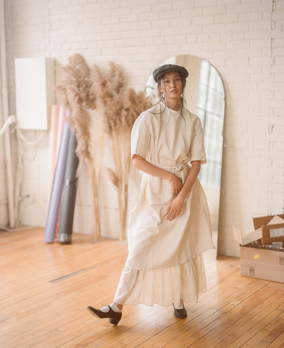 A woman in a vintage nursing uniform and cap stands on a wooden floor in a room with a mirror and dried plants.