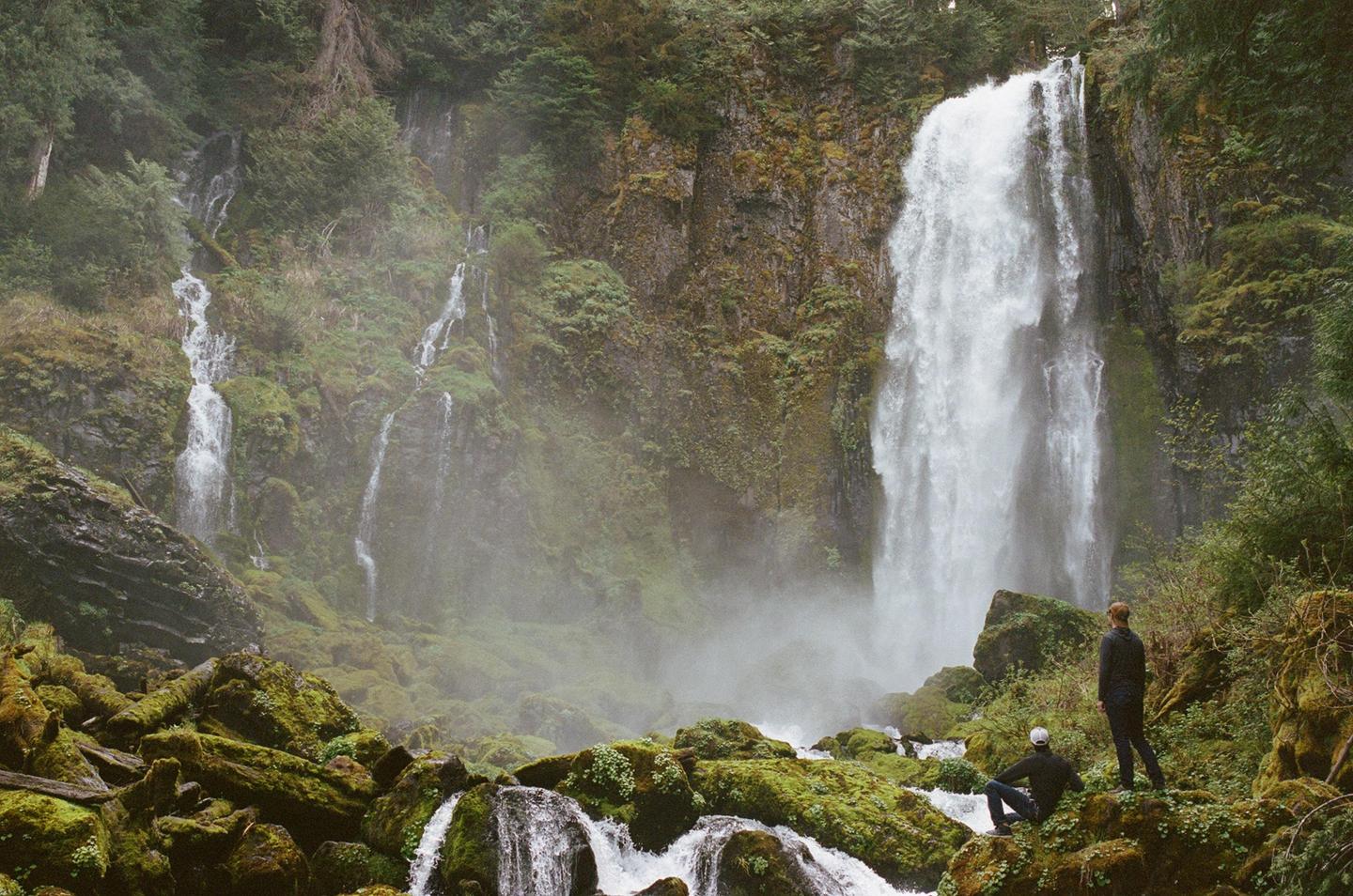 Two men stand and sit on mossy rocks looking at a large waterfall and smaller cascades in a lush, green forest.