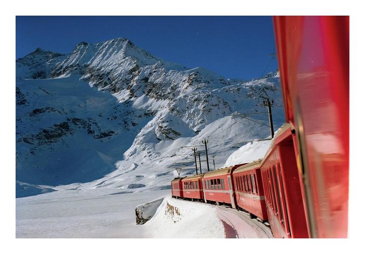 A red train travels along a snowy mountain landscape under a clear blue sky.