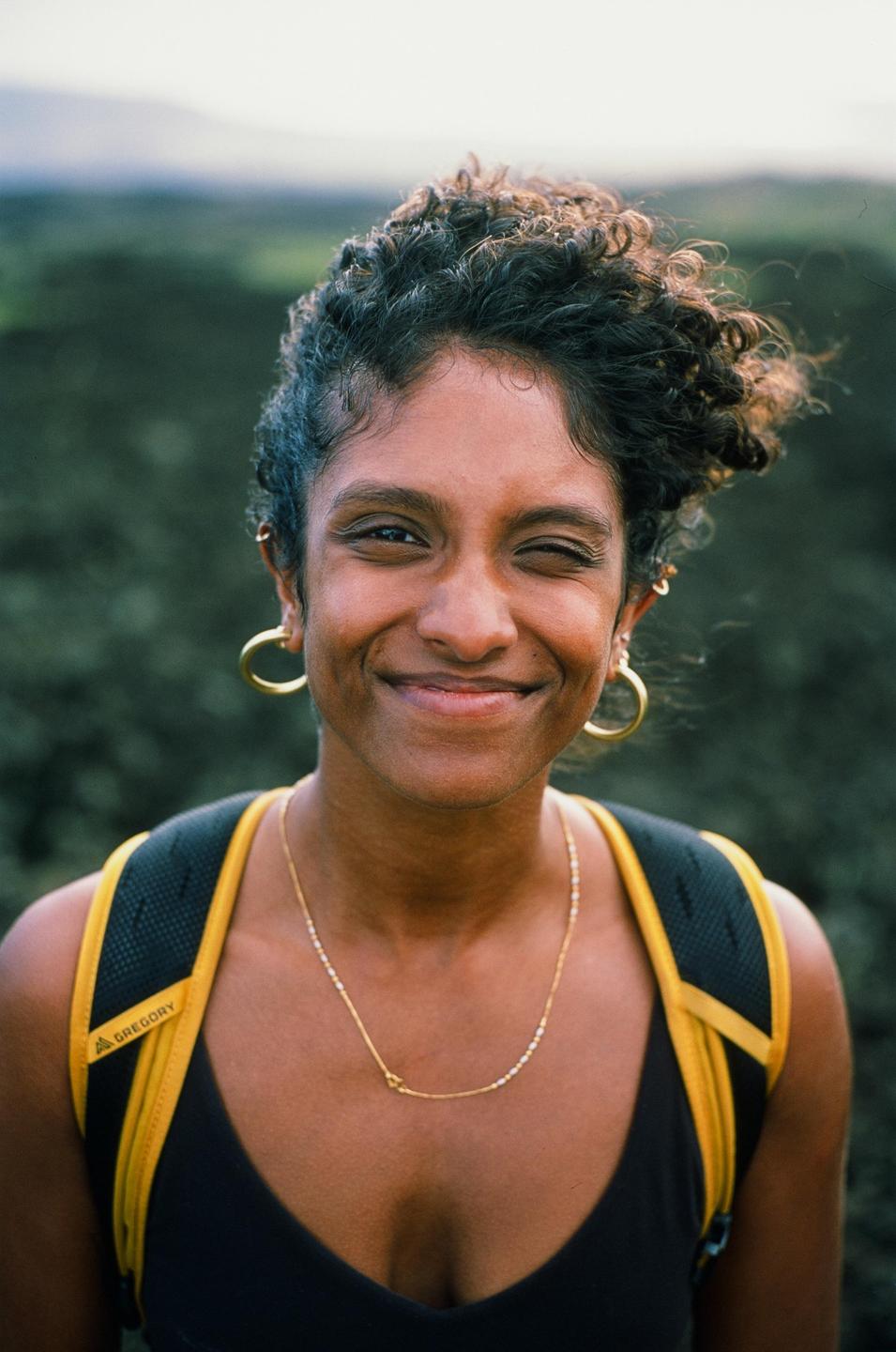 A woman with curly hair and a backpack smiles and winks at the viewer.