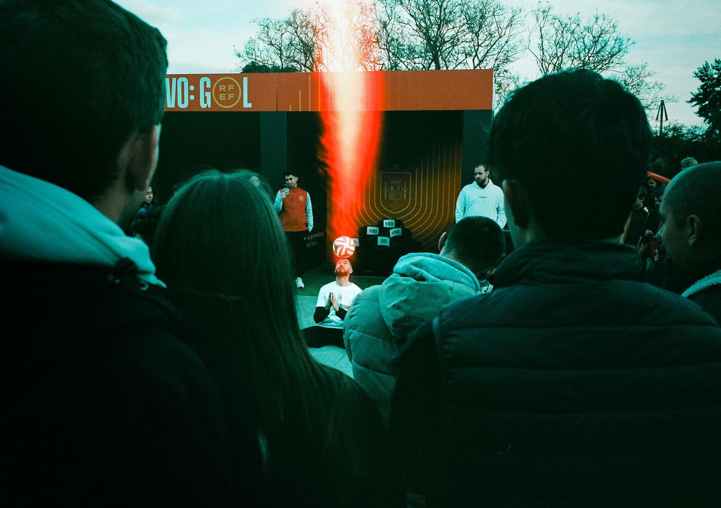 A man balances a soccer ball on his head while sitting in front of a screen with a red light leak.