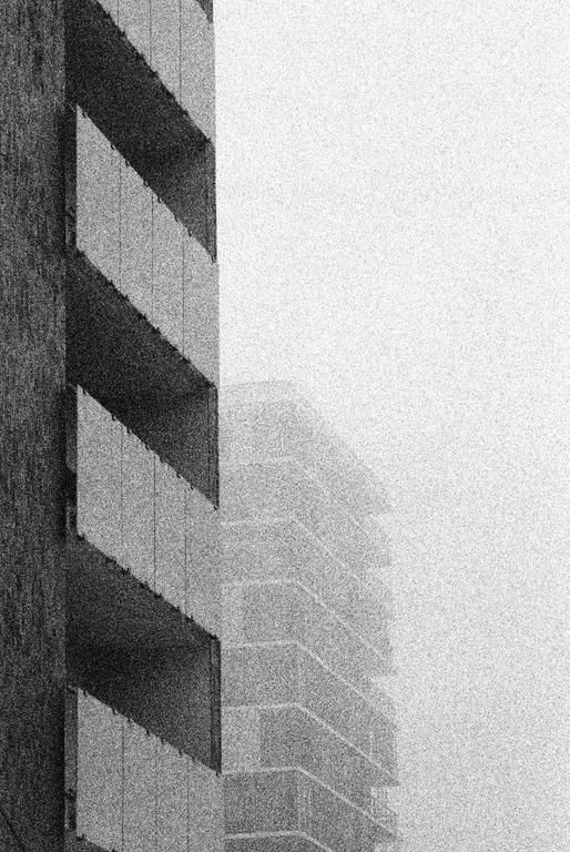 A tall building with balconies is partially obscured by fog, with another building faintly visible in the background.