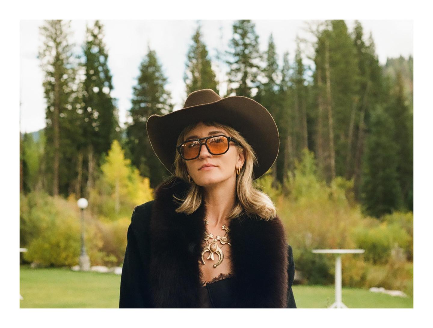 A woman wearing a cowboy hat and sunglasses stands in a grassy area with trees in the background.