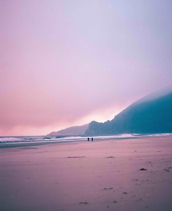 Two people stand on a wide beach with waves crashing near a tree-covered mountain under a pink and purple sky.