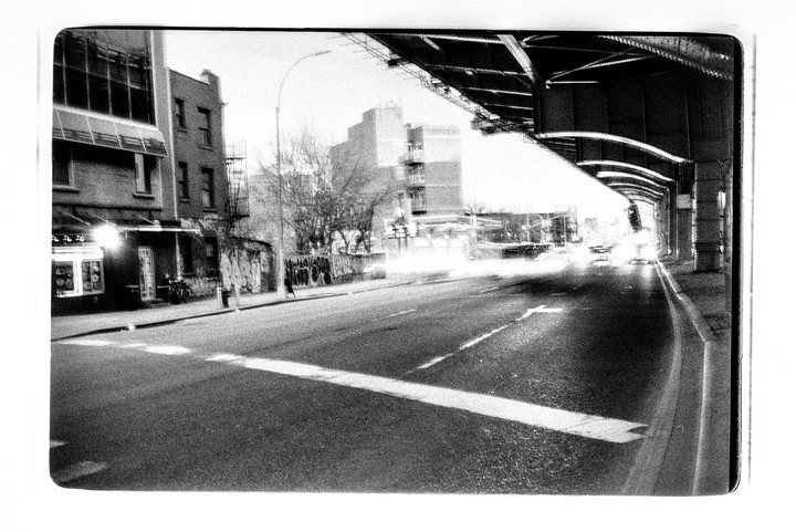 A street with light trails from vehicles passes under an elevated structure next to buildings with graffiti.