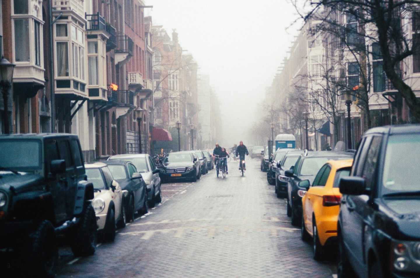 Two people ride bicycles down a foggy street lined with parked cars and buildings.