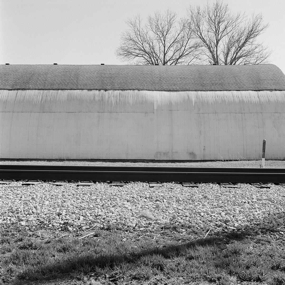A train track runs in front of a long building with a bare tree behind it.
