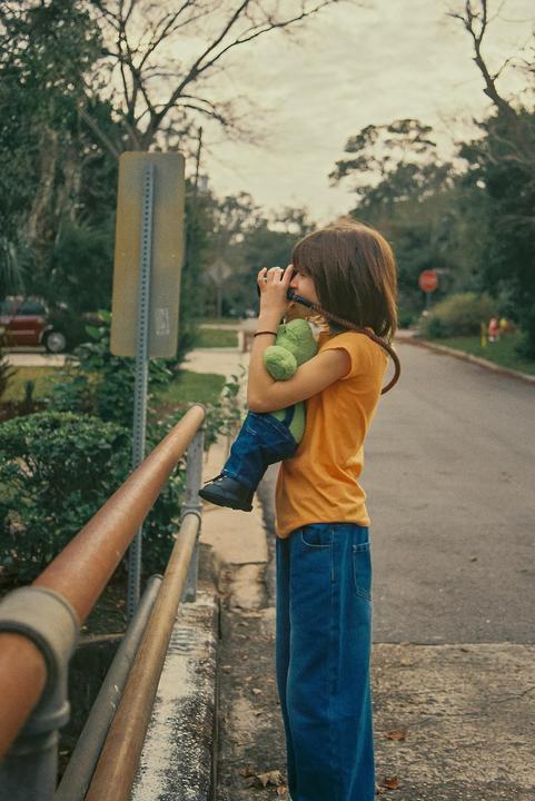 A child in a yellow shirt and blue jeans holds a green stuffed animal while looking through a camera on a street.