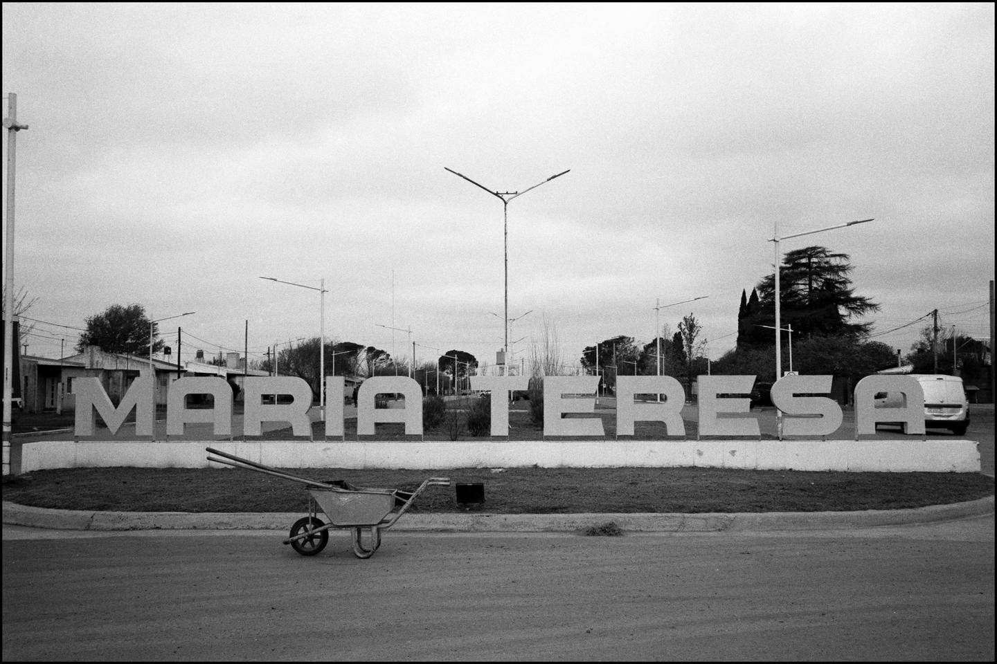A wheelbarrow sits on the road in front of a large sign that reads "MARIA TERESA" in a small town.