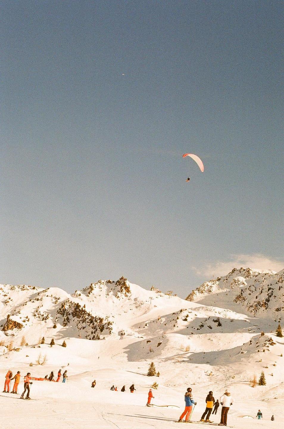 A paraglider flies over a snowy mountain landscape with skiers on the slopes below.