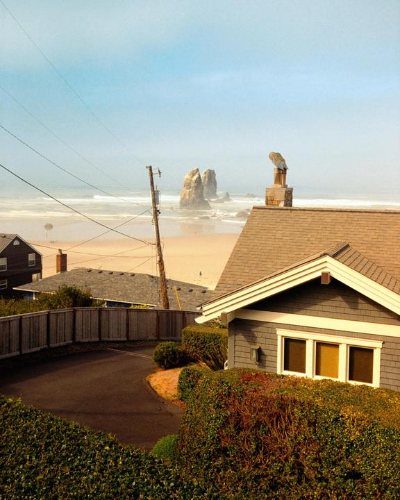 A house with a driveway and hedges overlooks a beach with large rock formations in the distance.