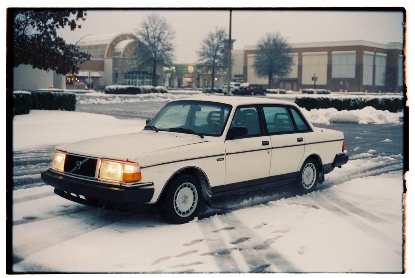 A white car is parked in a snowy parking lot with a shopping mall in the background.