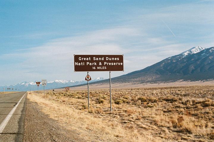 A brown sign for Great Sand Dunes National Park and Preserve stands beside a road with mountains in the background.