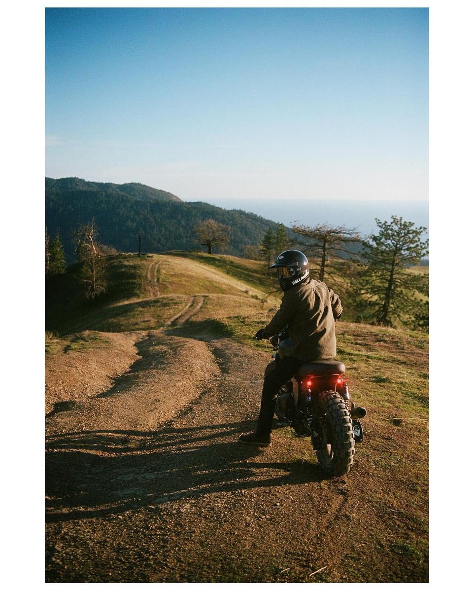 A man on a motorcycle looks back while riding on a dirt trail overlooking the ocean.