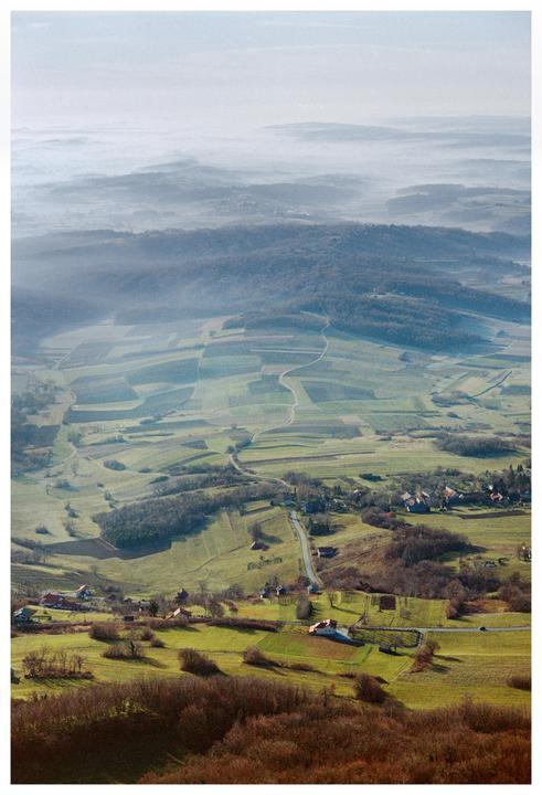 A winding road leads through a valley of fields and houses, with hazy hills in the distance.