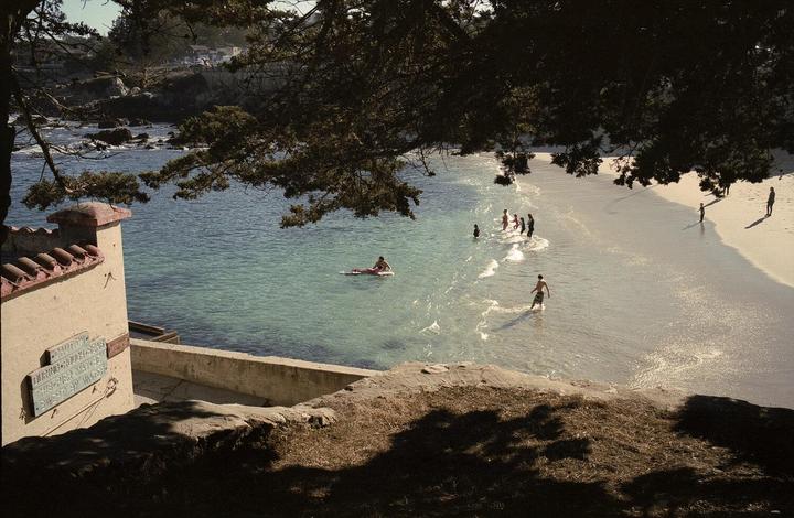 A group of people are swimming and wading in the ocean next to a sandy beach, with a person on a surfboard in the water and a building with a red tile roof on the left.