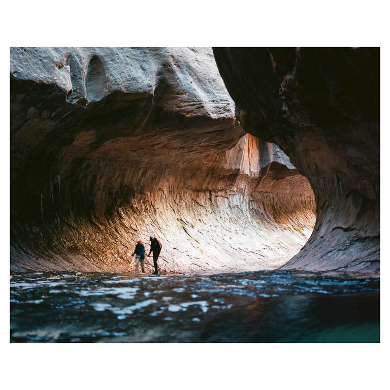 Two people stand in a narrow canyon with water in the foreground.
