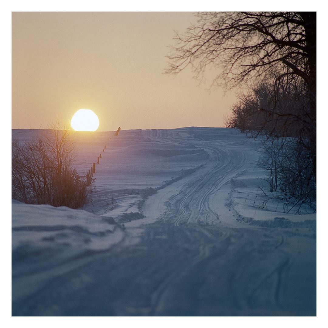 The sun rises over a snow-covered hill with a path and trees.