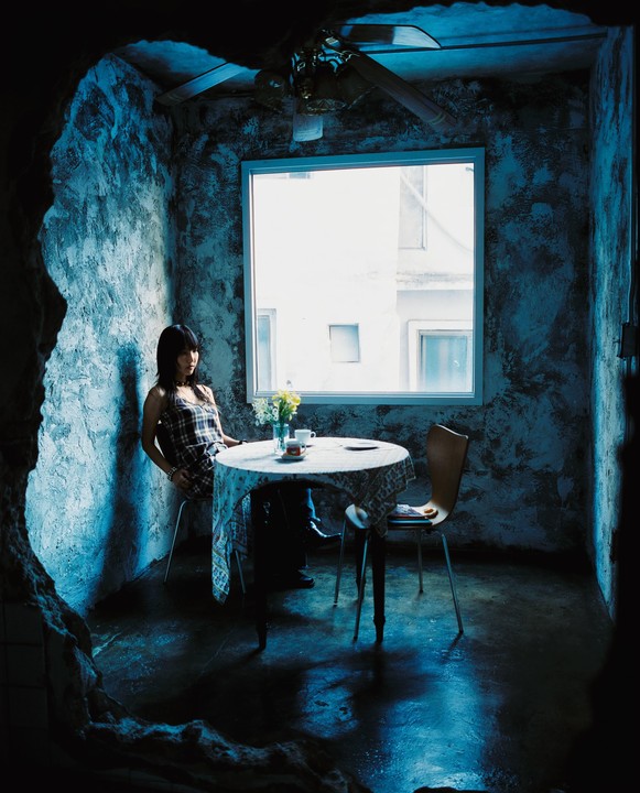 A woman sits at a table with a cup of coffee and flowers next to a window in a dimly lit room.