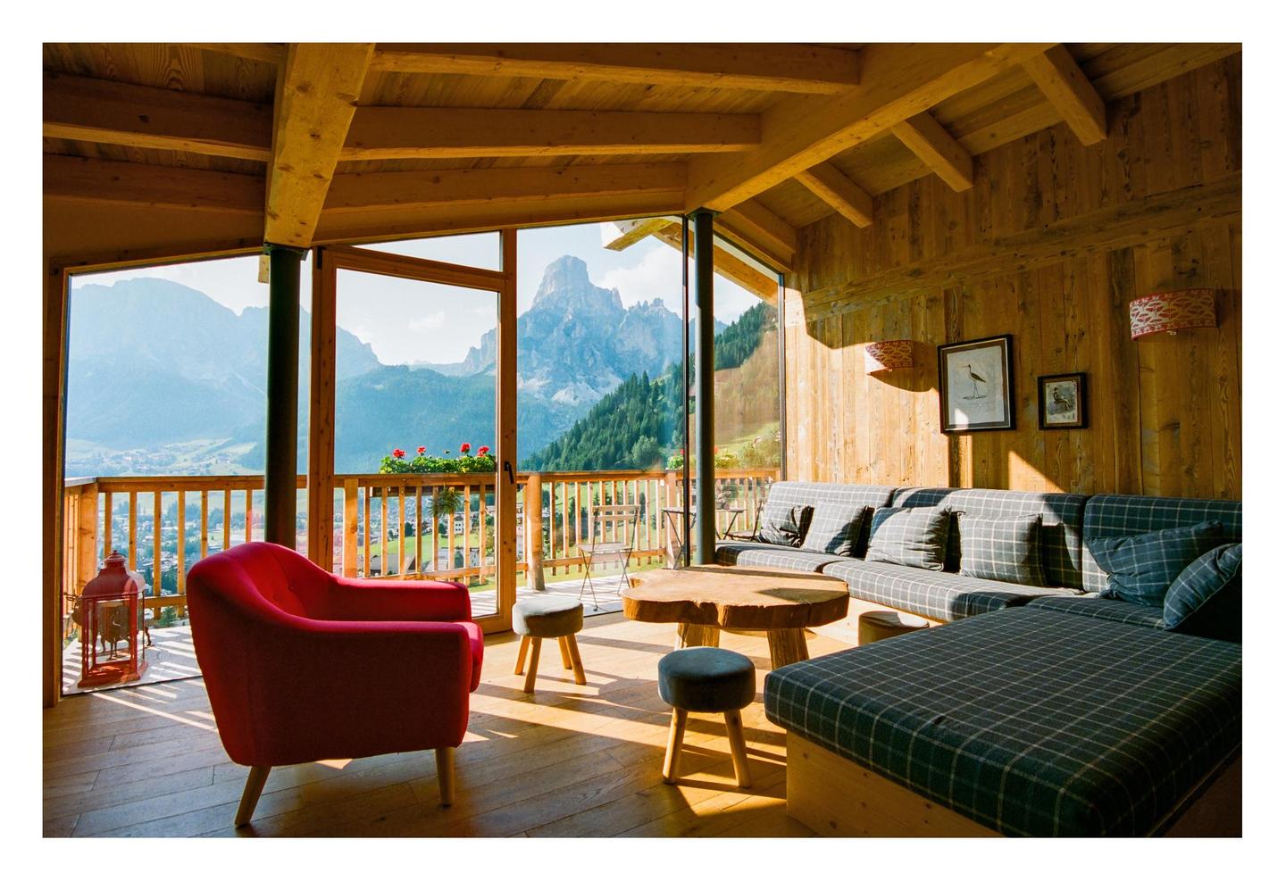 A cozy room with wooden walls and ceiling features a red armchair, a plaid sofa, and a wooden coffee table, all bathed in sunlight streaming through large windows that offer a panoramic view of mountains and a valley.