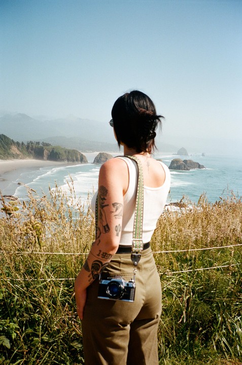A woman with tattoos on her arm stands with her back to the viewer, looking out at a coastline with rock formations in the water.