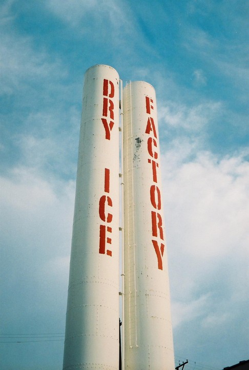 Two white industrial silos with red lettering that reads "DRY ICE FACTORY" stand against a blue sky with clouds.