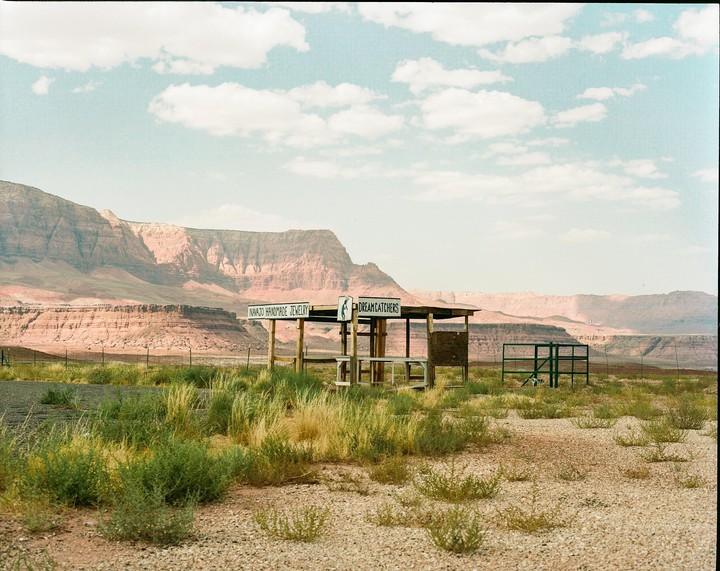 A small wooden stand with signs for "Navajo Handmade Jewelry" and "Dreamcatchers" sits in a desert landscape with mountains in the background.