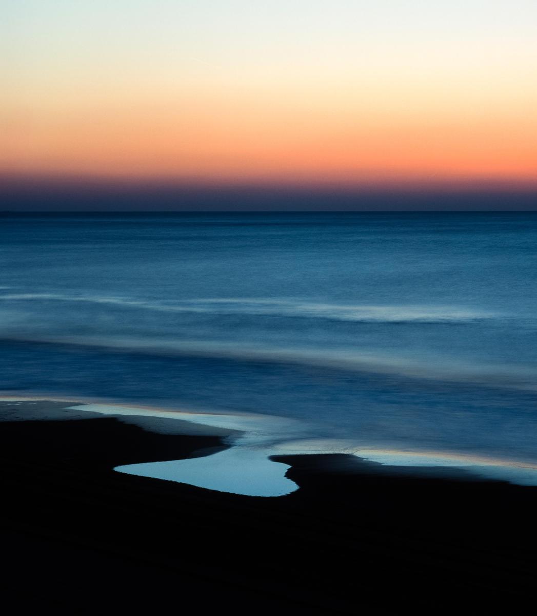 The ocean meets a dark beach with a small pool of water under a colorful sunset sky.