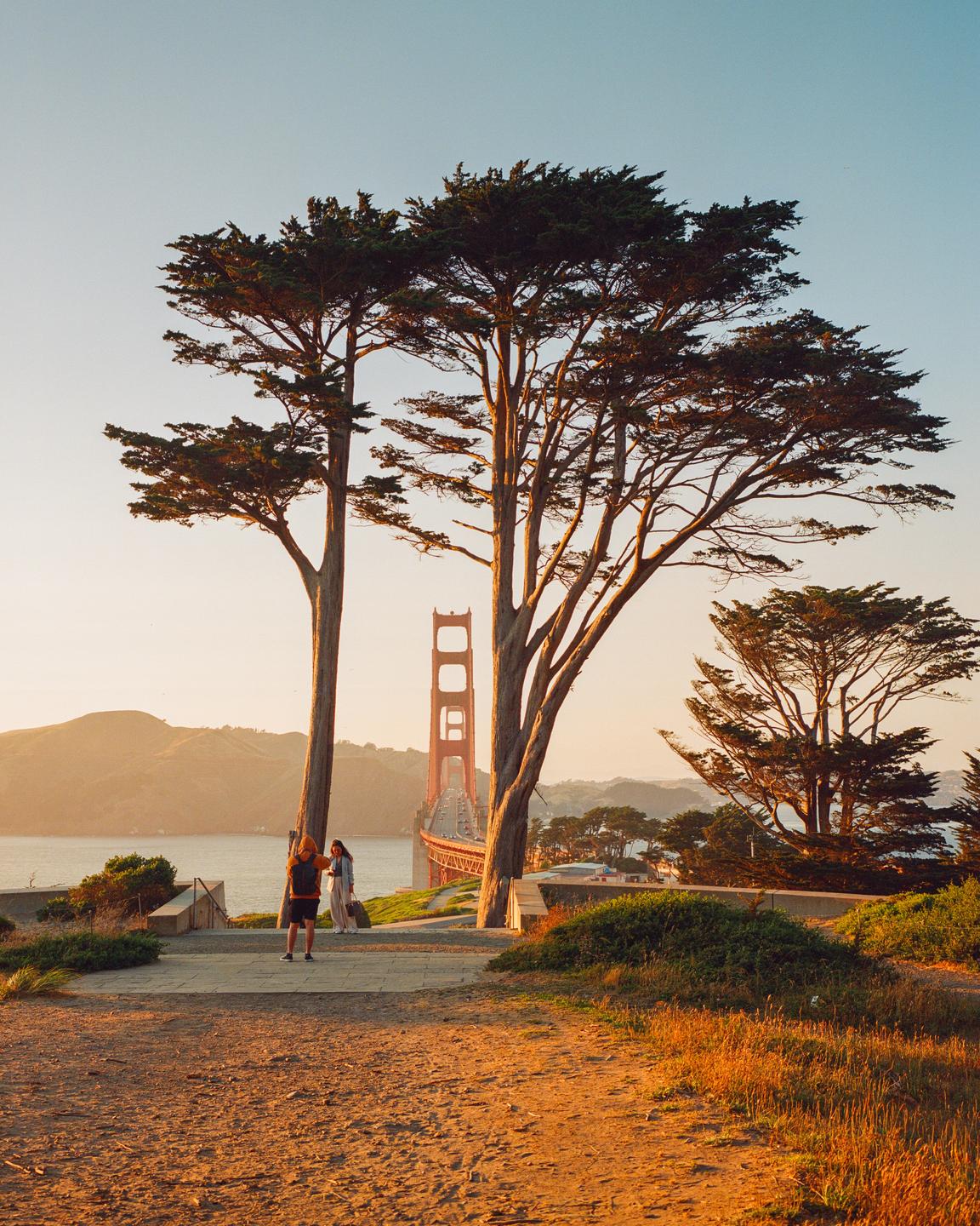 Two people stand on a path overlooking the Golden Gate Bridge at golden hour, framed by two large trees.