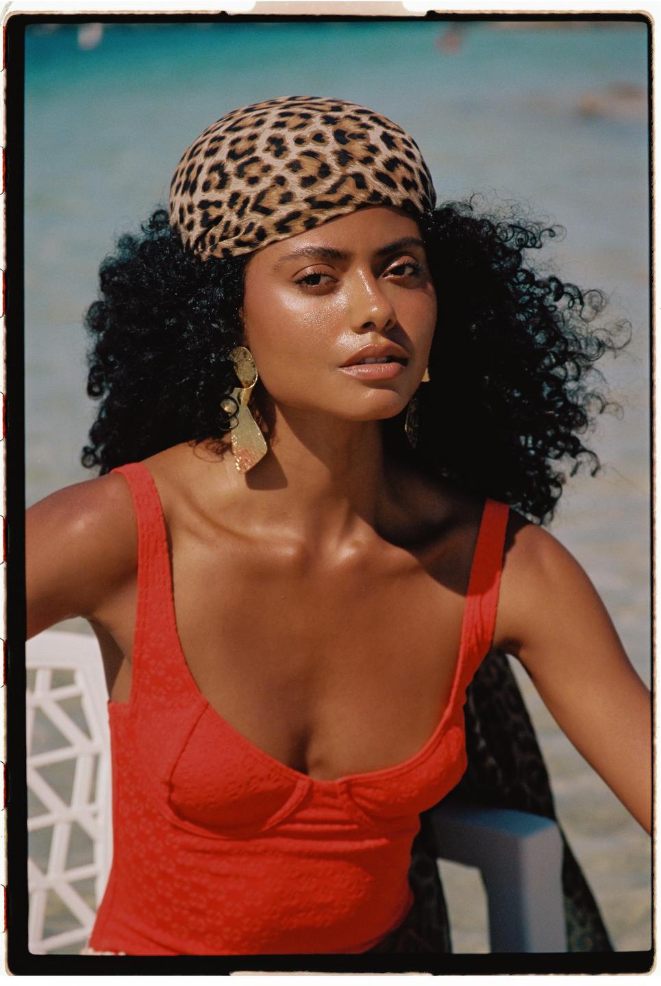 A woman with curly hair and a leopard print headscarf wears a red top and large gold earrings.