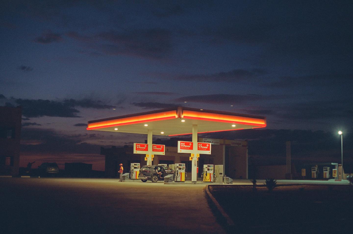 A gas station with a car and a man standing next to a pump is illuminated at dusk.