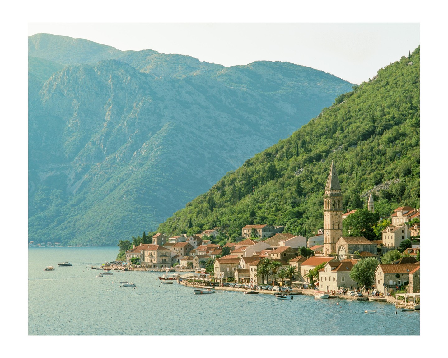 A small town with a prominent bell tower sits on the edge of a body of water, backed by a large, green mountain.