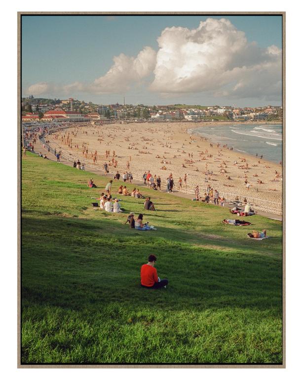 A man sits on a grassy hill overlooking a crowded beach with people sunbathing and swimming.