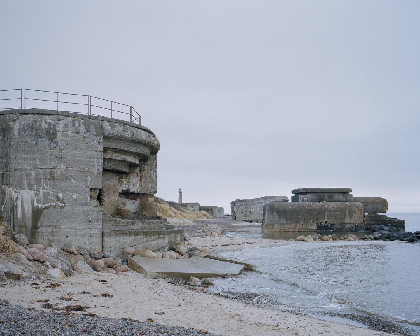 Concrete bunkers line a sandy beach with a lighthouse in the distance under a pale sky.