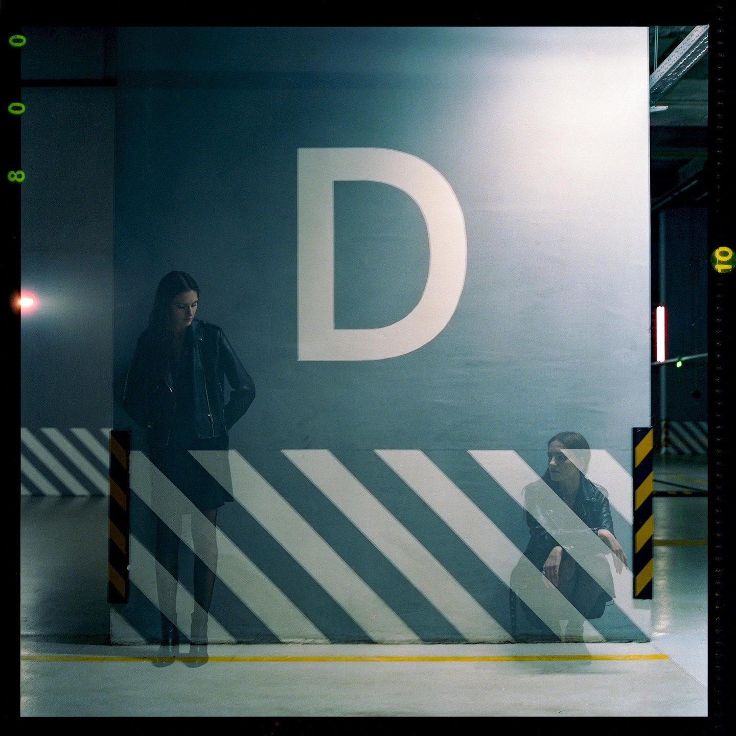 A double exposure shows a woman standing and a woman crouching in a parking garage with a large letter D on the wall.