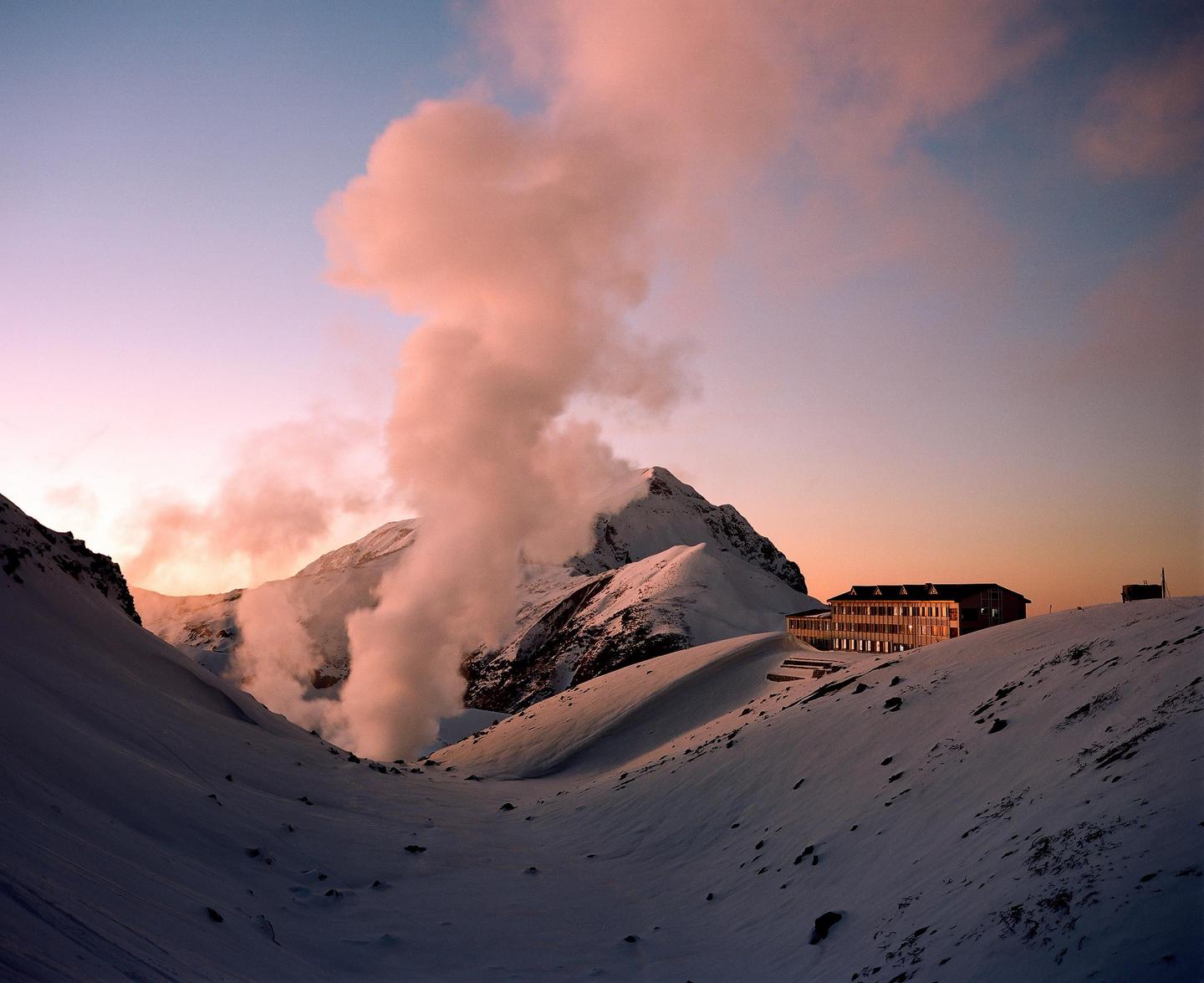 A building sits on a snow-covered mountain with steam rising from a valley at sunset.